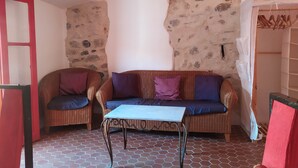 A room with a stone wall, two wicker sofas with cushions, a small table, and a tiled floor. - Attractive, Renovated Village House in Traffic-free Street (Estagel)