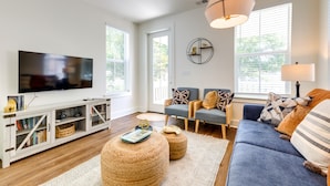 Ein modernes Wohnzimmer mit Flachbildfernseher, einer Couch, zwei Sesseln, einem Couchtisch, Hocker und einem Teppich mit Muster. Eine Wandleuchte ist angebracht.