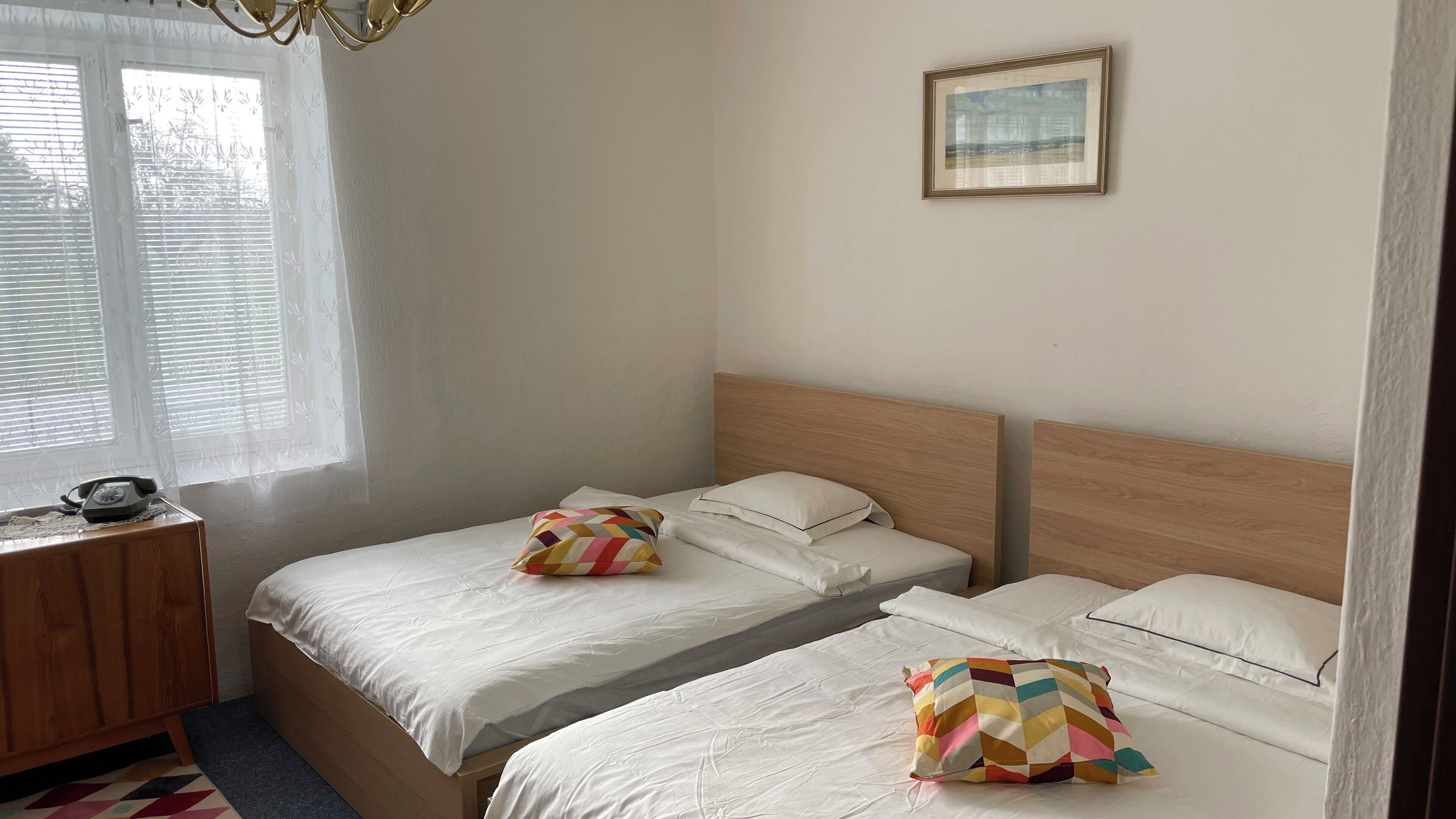 A hotel room with two single beds, a wooden headboard, a bedside table with a telephone, and a window with blinds.