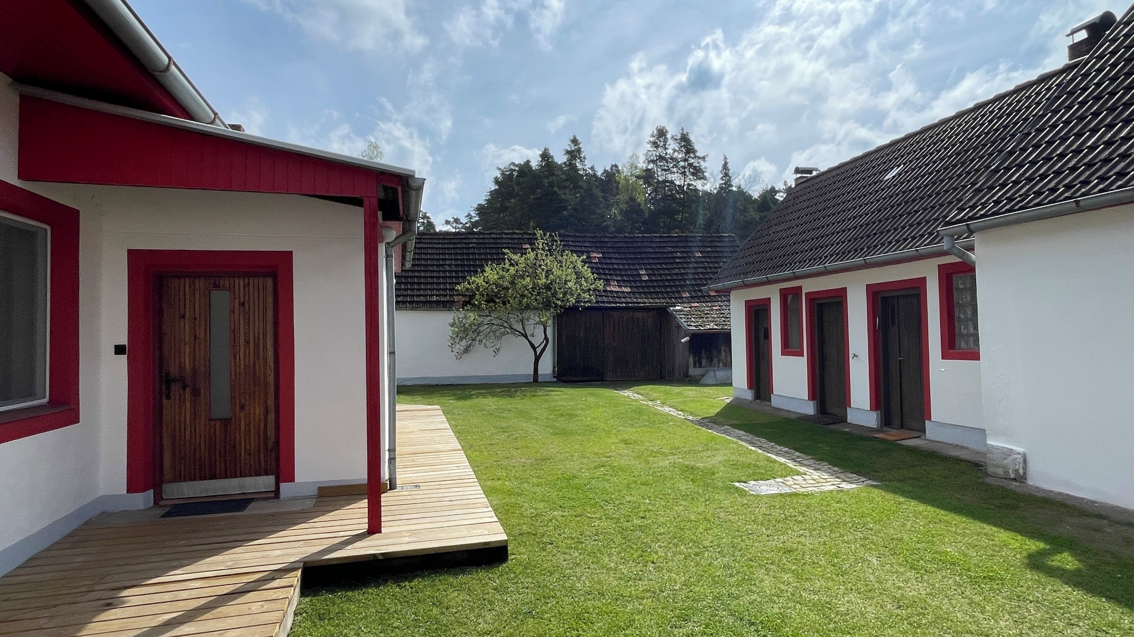 A courtyard with two white buildings with red trim, a wooden deck, and a small tree.