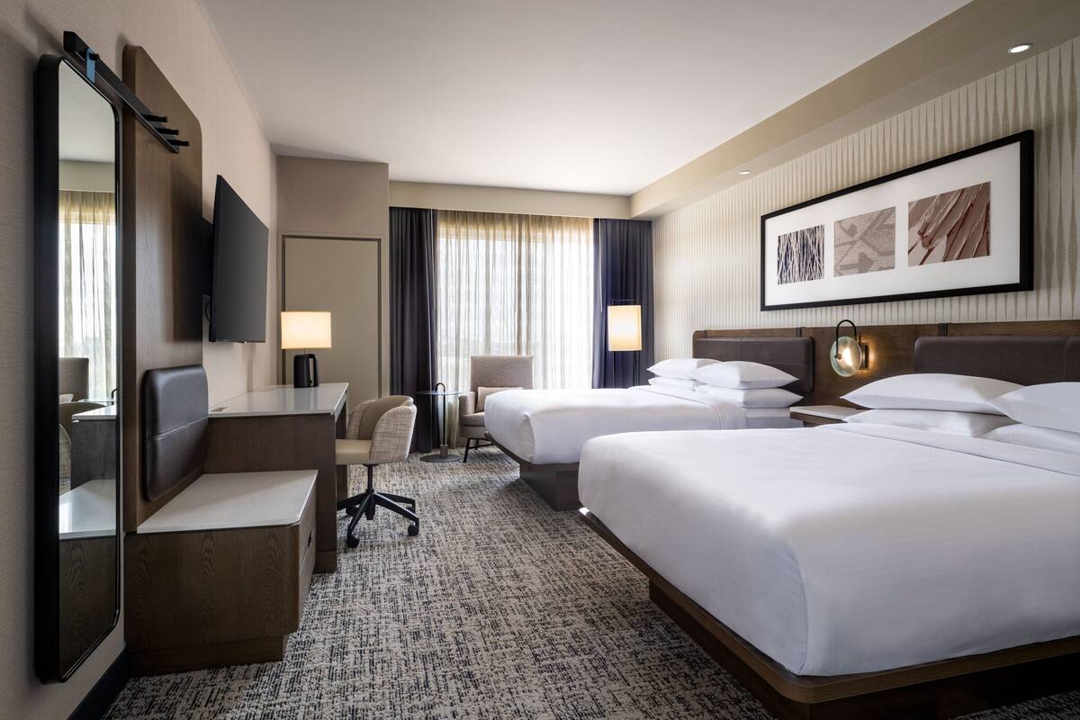 A hotel room with two beds, a desk, a chair, a flat-screen TV, a lamp, and a framed picture on the wall.