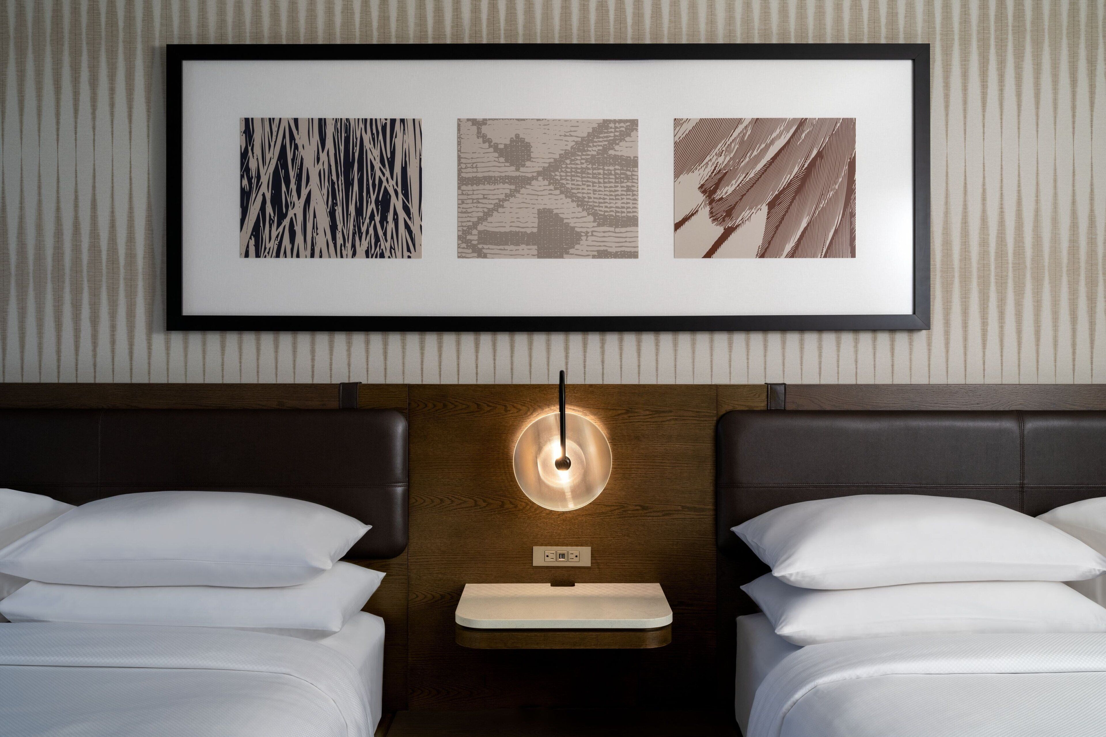 Een hotelkamer met twee bedden, een nachtkastje, een lijst met kunstwerk erboven en een wandlamp.