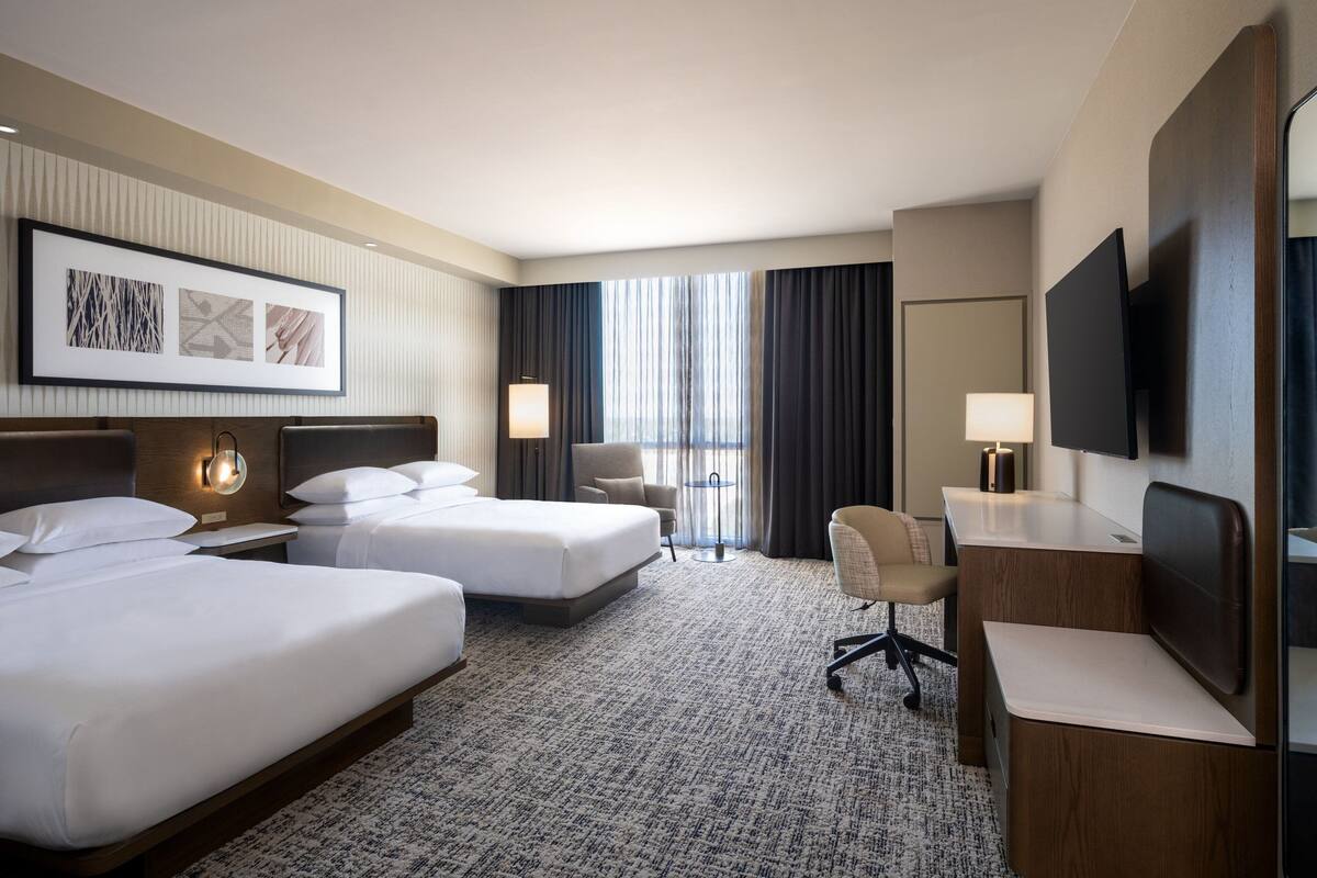 A hotel room with two beds, a desk, a chair, a TV, and a large window with curtains.