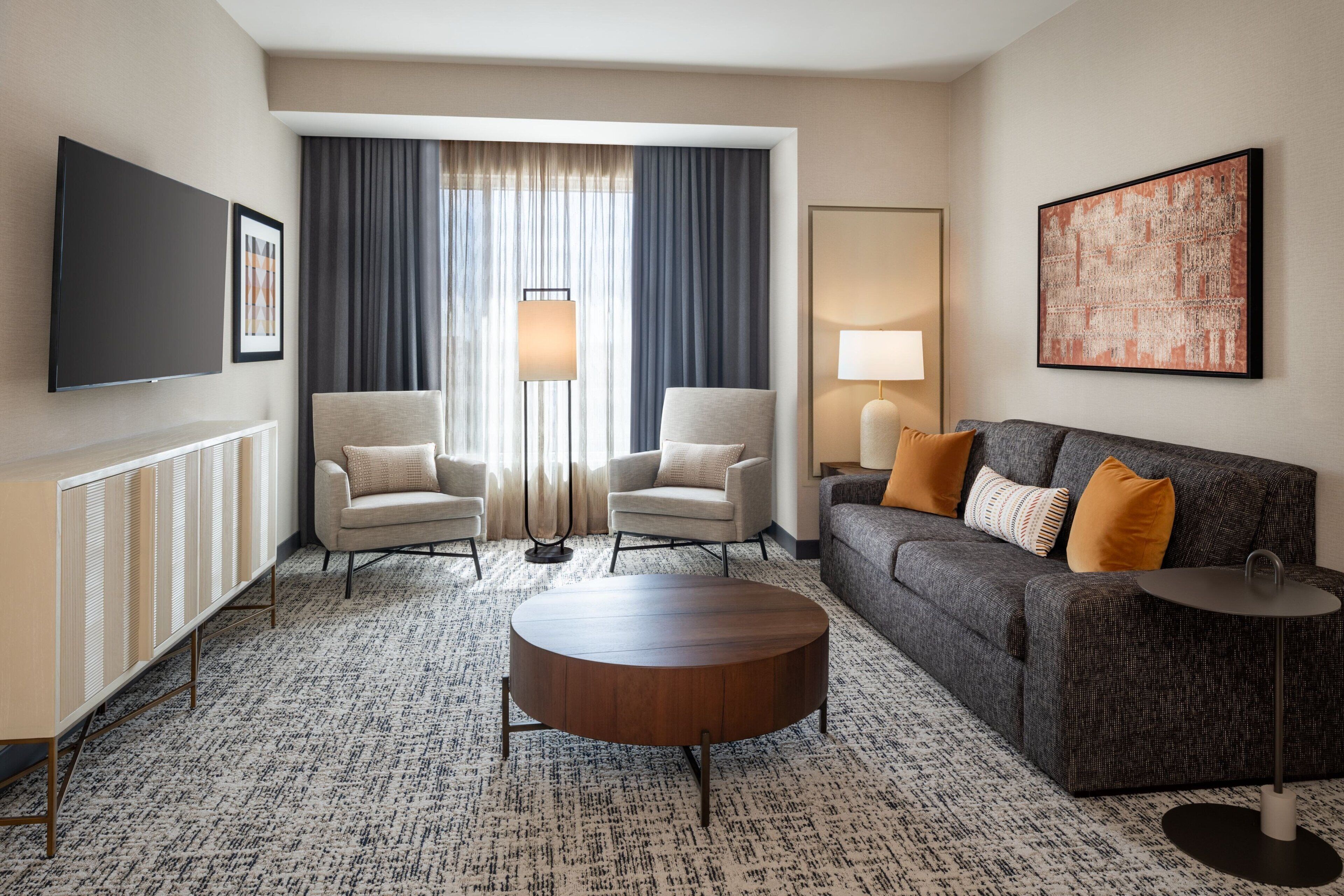 A modern hotel room with a large window, two armchairs, a sofa, a coffee table, and a flat-screen TV.