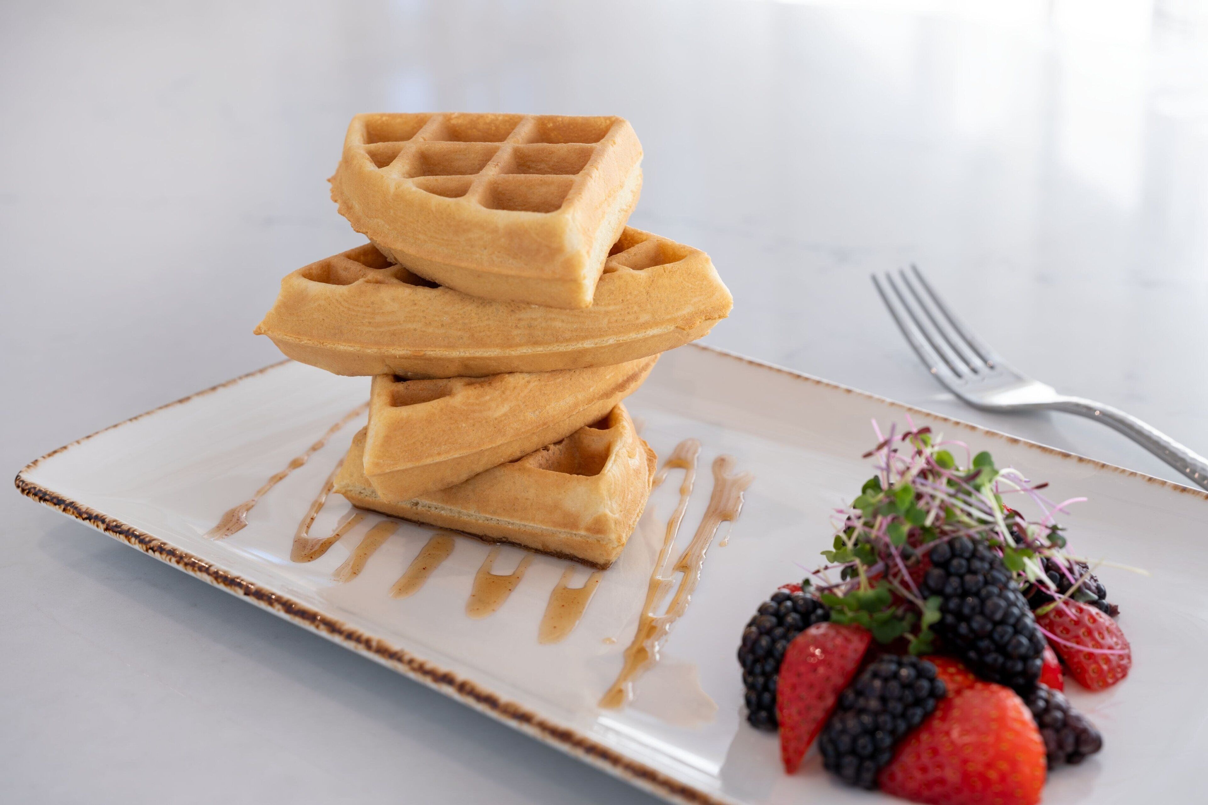 A stack of waffles on a plate with a side of mixed berries and a drizzle of syrup.