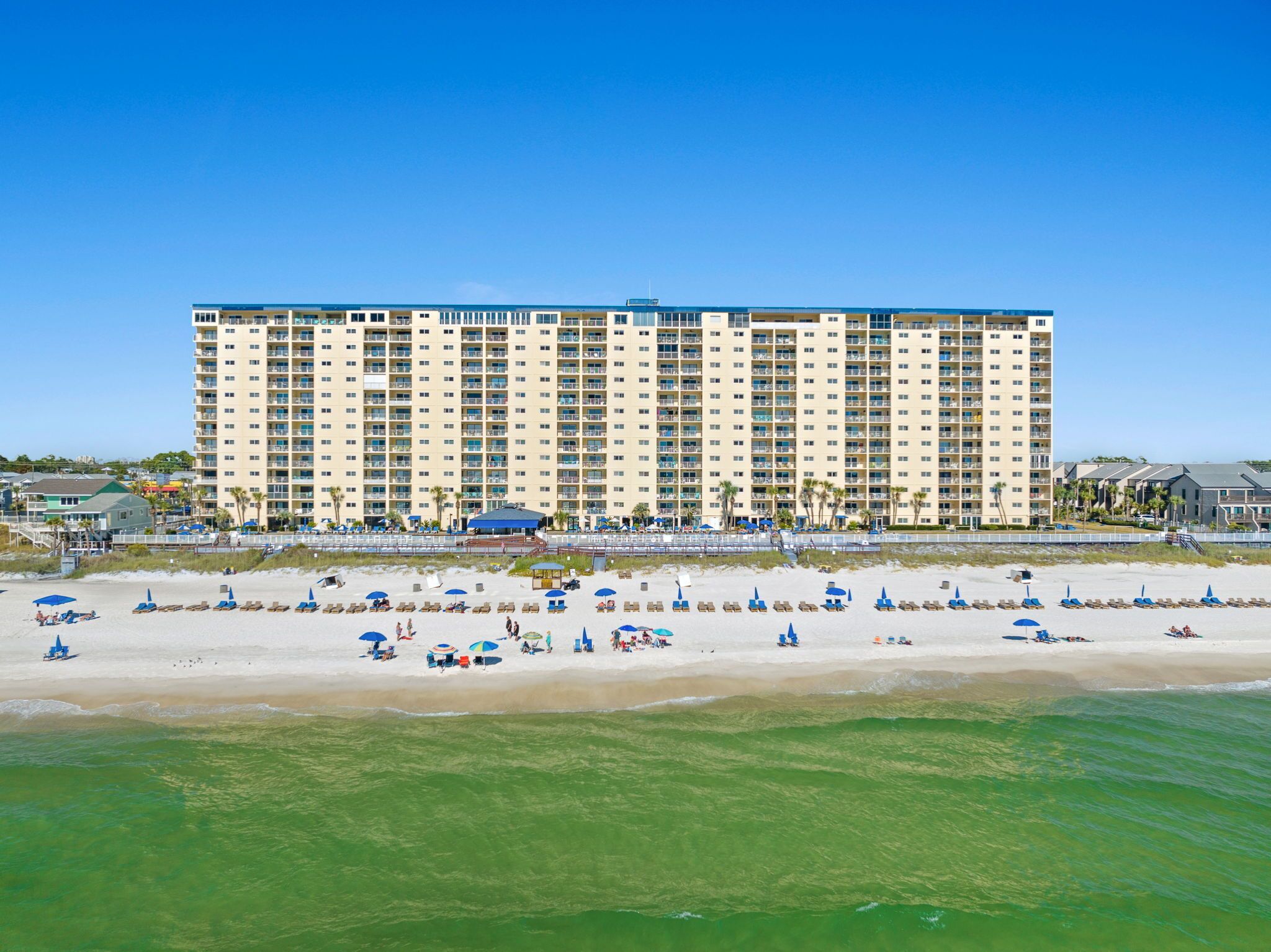 Een groot hotel met meerdere verdiepingen en balkons, een strand met ligbedden en parasols, en een helderblauwe lucht.