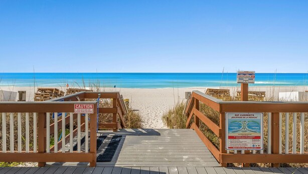 Een houten looppad dat naar een strand leidt, met helderblauw water, een waarschuwingsbord en een bord met waarschuwing voor stroming.