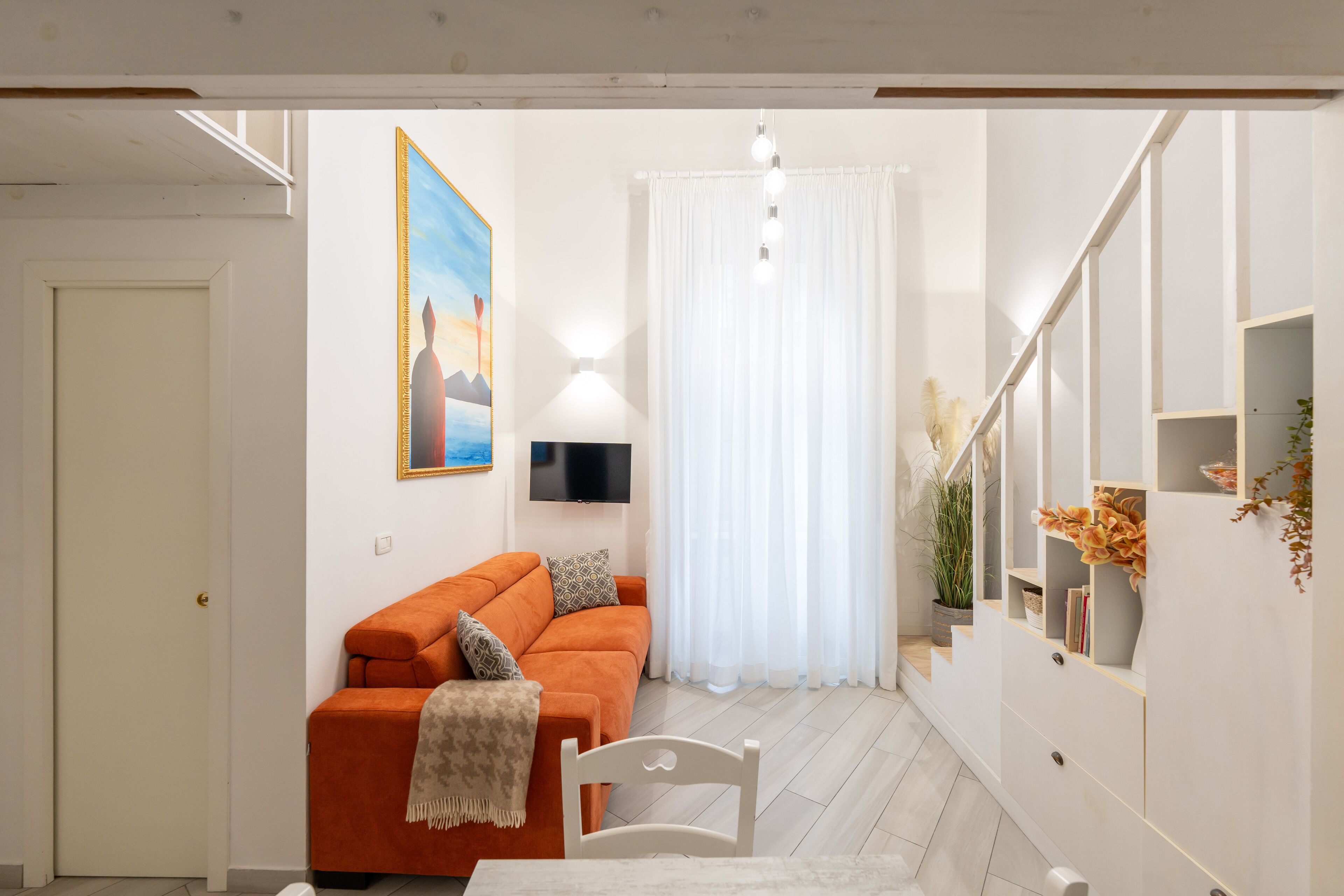 A compact living space with a sofa, a dining table, a TV, and a staircase with a plant.