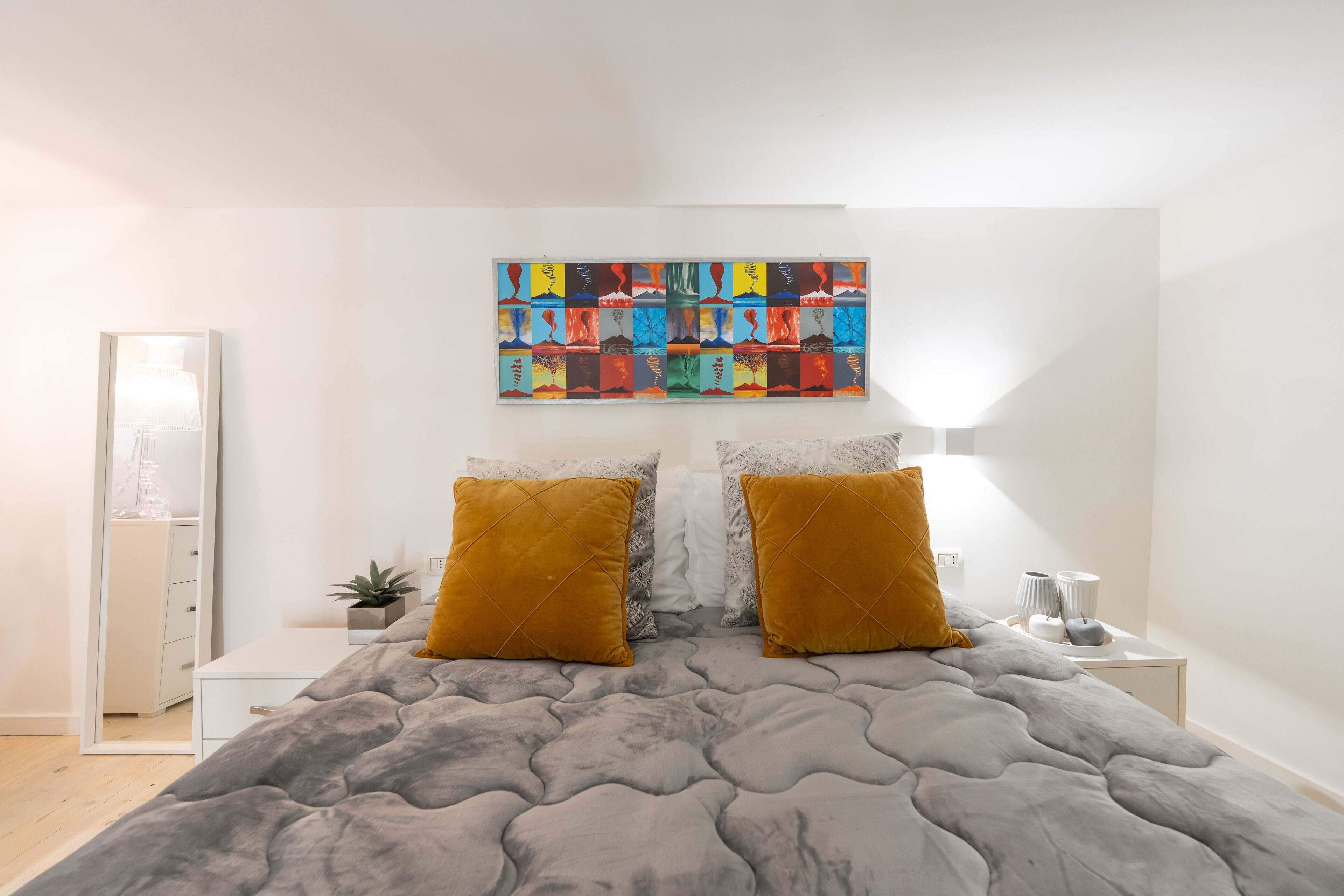 A bedroom with a grey quilted bedspread, two mustard pillows, a colorful abstract painting above the bed, a bedside table with a lamp, and a small plant.