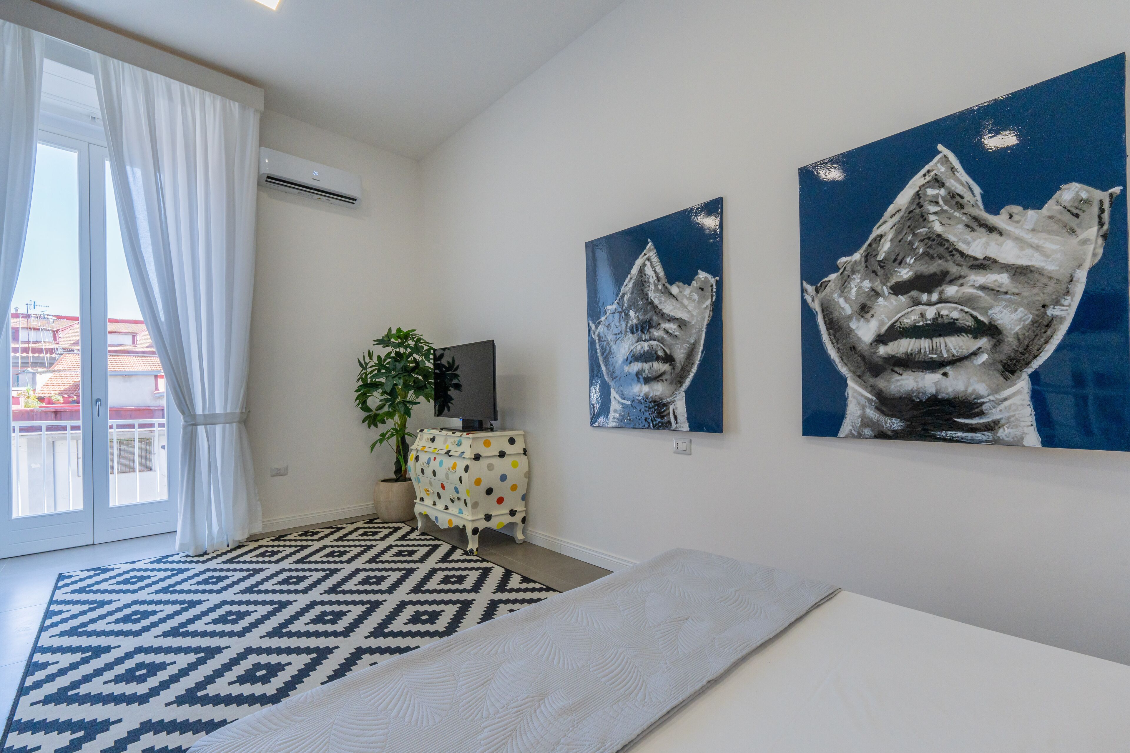 A modern bedroom with a large window, a patterned rug, a white mattress, a polka-dotted dresser, and two abstract paintings on the wall.