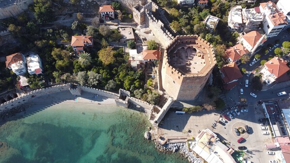An aerial view of a coastal town with a prominent castle, clear turquoise waters, and a mix of residential and commercial buildings. - AGORA APART OTEL (Alanya)