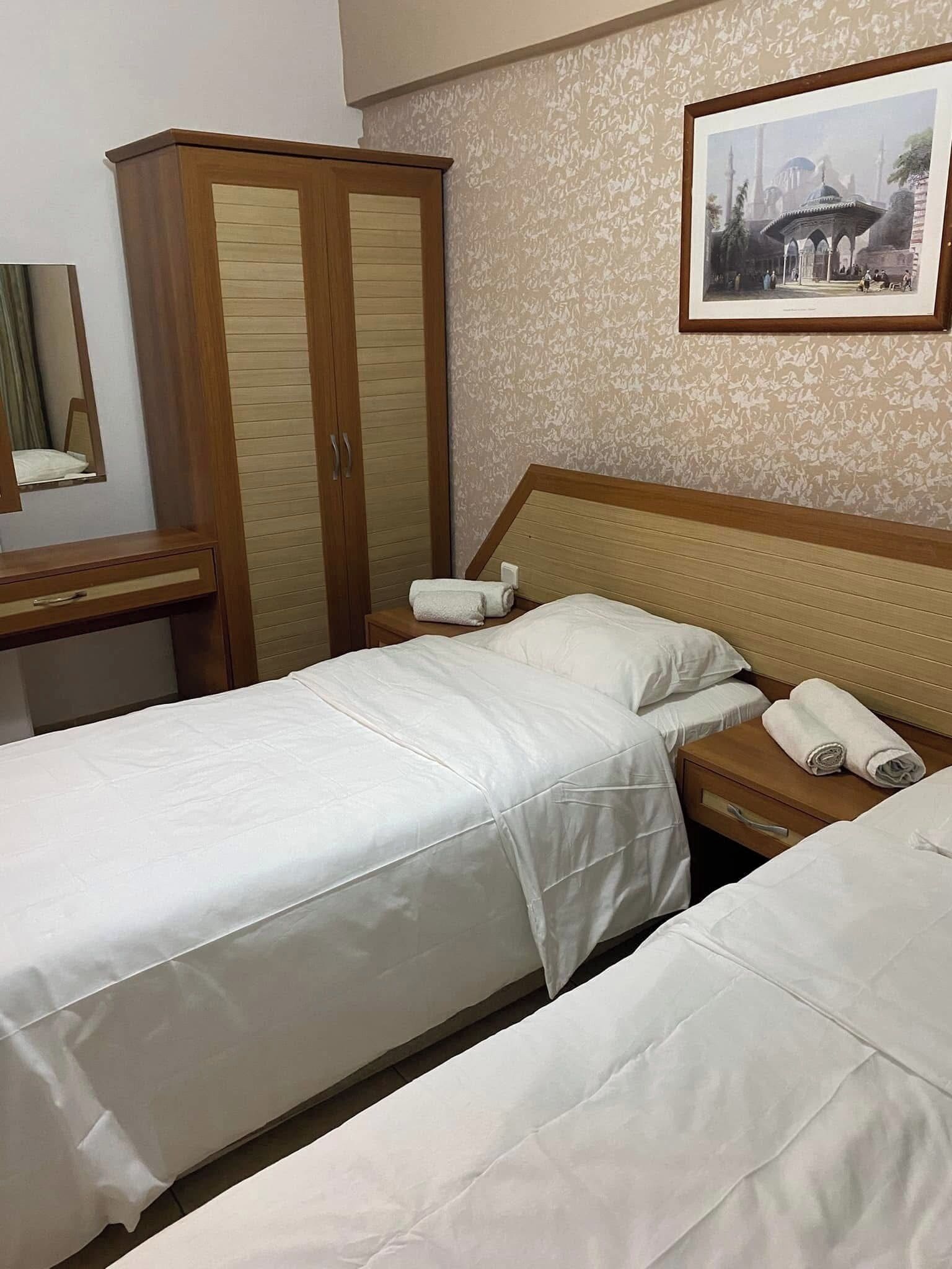 Habitación de hotel con dos camas individuales, un armario de madera, una mesita de noche con espejo y un cuadro en la pared.