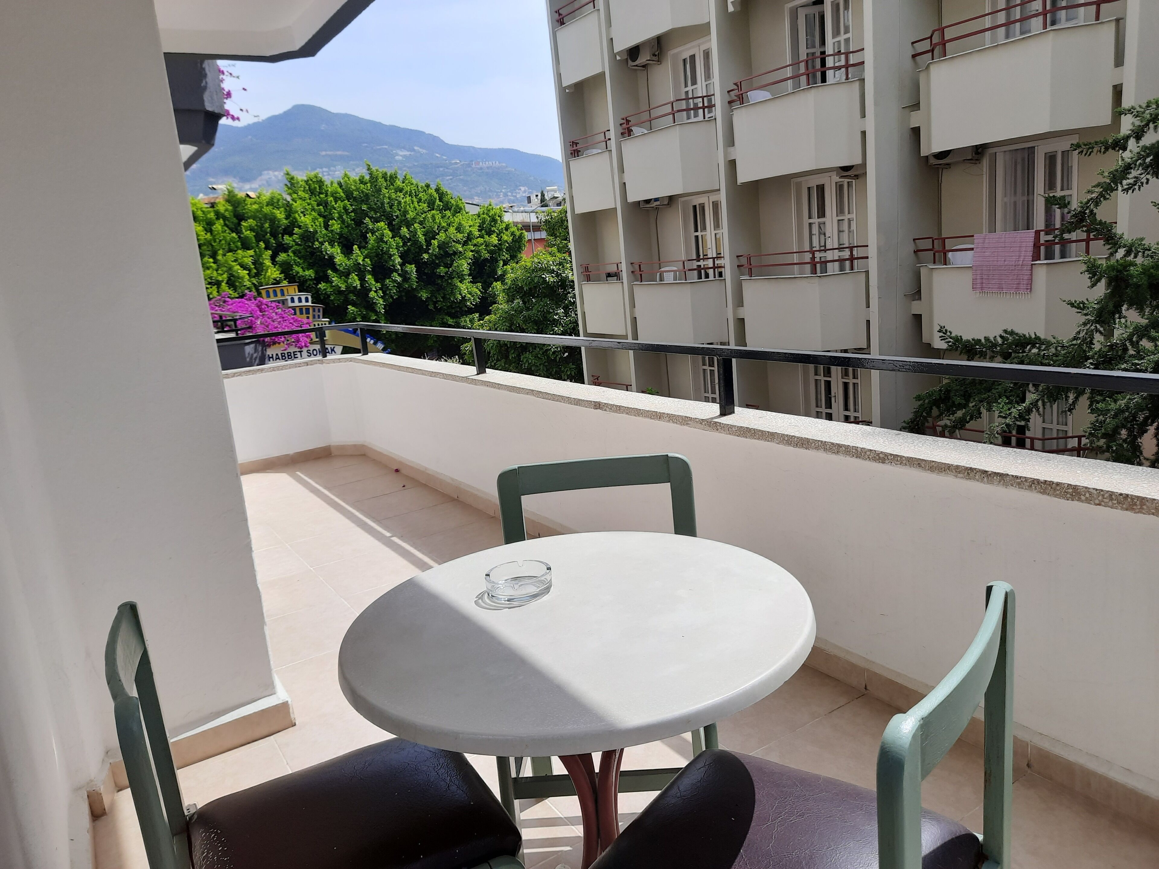 A balcony with a round table and two chairs, a view of a mountain, and a building with balconies in the background.