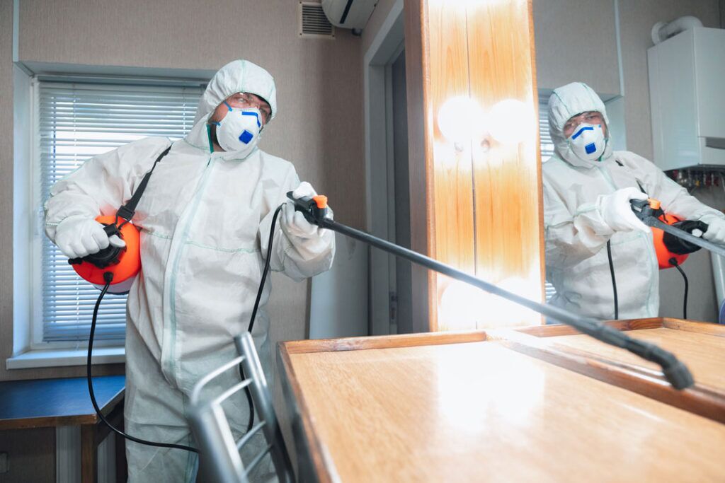 Una persona con traje de protección usando un pulverizador en una habitación con una mesa de madera y una ventana con persianas.