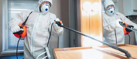 Una persona con traje de protección usando un pulverizador en una habitación con una mesa de madera y una ventana con persianas.