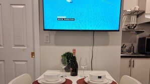 Un comedor con una mesa blanca para dos personas, un televisor mostrando una escena marina azul, y una cocina al fondo.