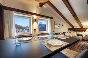 Uma sala de jantar com mesa posta para quatro pessoas, duas janelas com vista para as montanhas, um sofá e um abajur.
