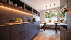 A modern kitchen with built-in appliances, a dining area with a bench, and a large window with a view of trees. - 191A Apartments Elmarino (Ortisei)