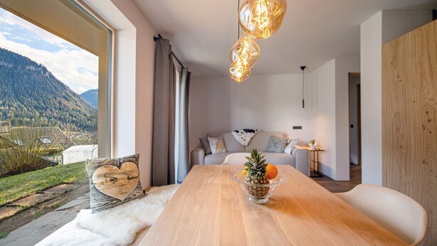 A modern dining area with a wooden table, a bowl of fruit, and a view of a mountainous landscape through large windows. - 191A Apartments Elmarino (Ortisei)