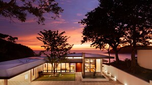 A modern house with a large glass facade, a covered patio, and a view of the sunset over the ocean. - Brand New Beach front 5 bedroom house steps from the beach  (San Juan del Sur)