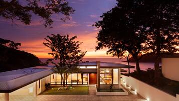 A modern house with a large glass facade, a covered patio, and a view of the sunset over the ocean.