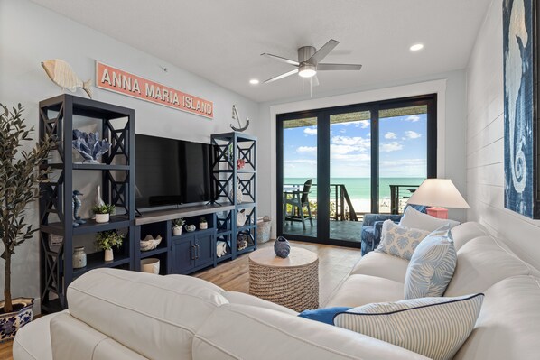 A living room with a large flat-screen TV, a sofa, a coffee table, and a bookshelf. There is a sign that reads "Anna Maria Island" and a view of the ocean through large windows. - The Gulf Front- 2 bedroom beachfront condo with incredible views (Holmes Beach)