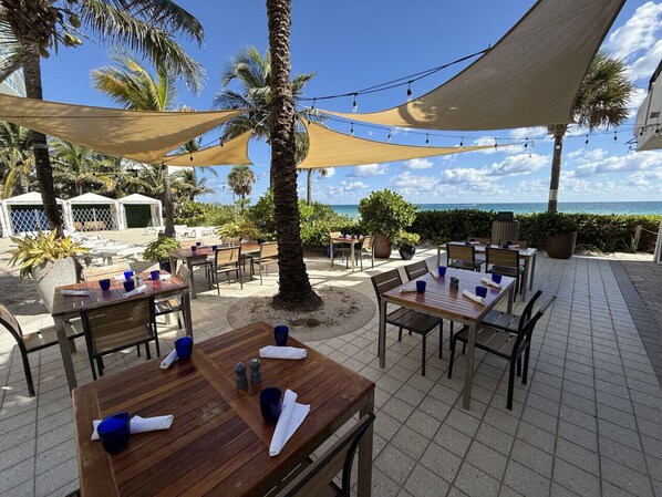 An outdoor dining area with tables and chairs, palm trees, and a view of the ocean.