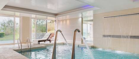 A modern indoor pool with clear water, metal ladders, and a lounge area with a chair and a table.