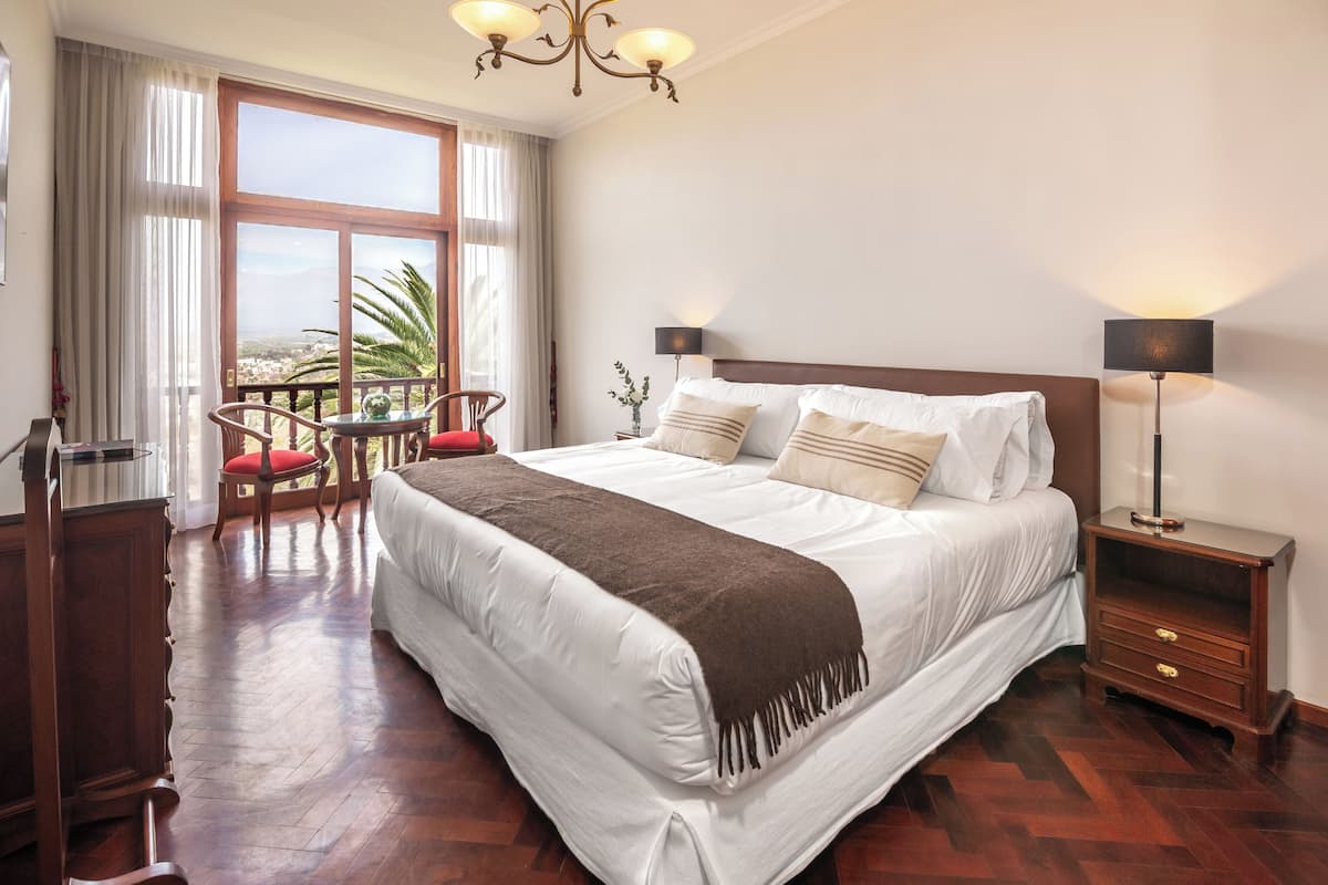 Une chambre d’hôtel avec un grand lit, un plancher en bois, un balcon avec vue, un bureau avec une chaise et une table de chevet avec une lampe.