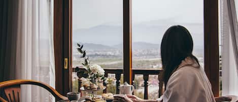 A dining table with a breakfast setting, including a pitcher, cups, and a variety of pastries, viewed from a balcony with a scenic mountain backdrop.