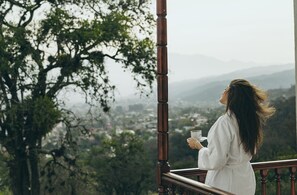 Una persona parada en un balcón con barandilla, sosteniendo una taza, con vista a un paisaje urbano que incluye árboles y montañas al fondo.