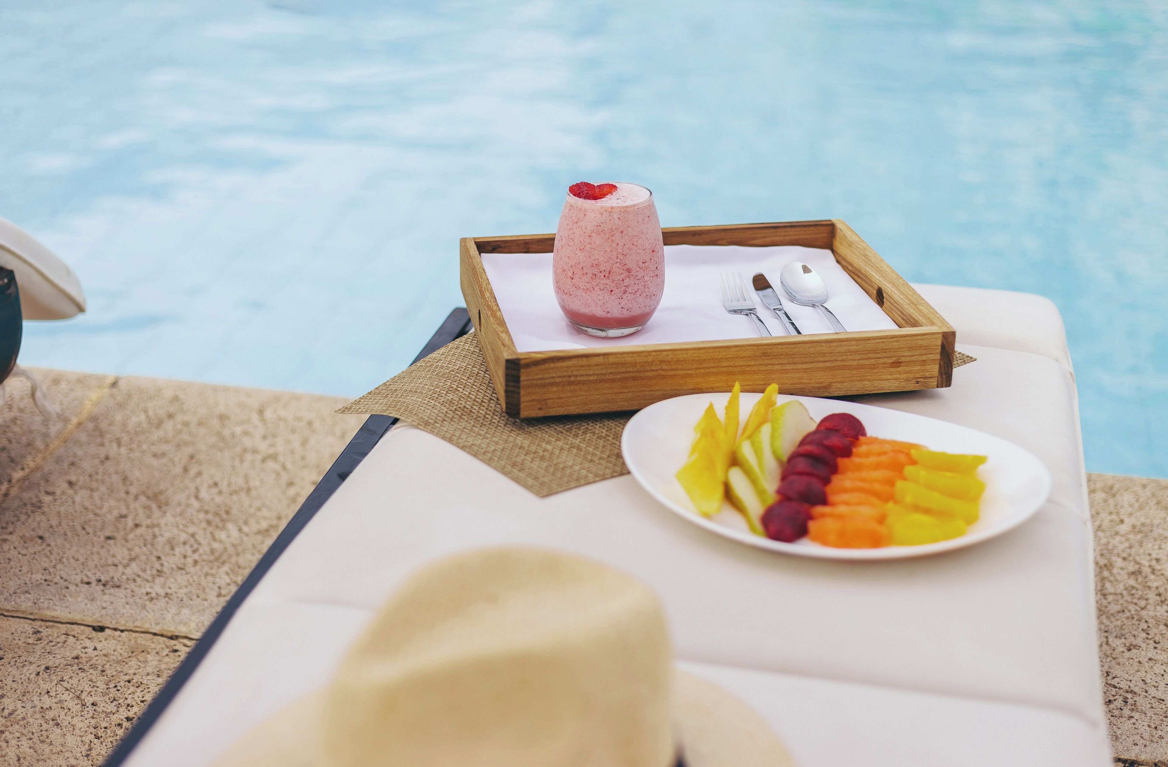 A tray with a fruit salad and a smoothie by a poolside lounge chair.