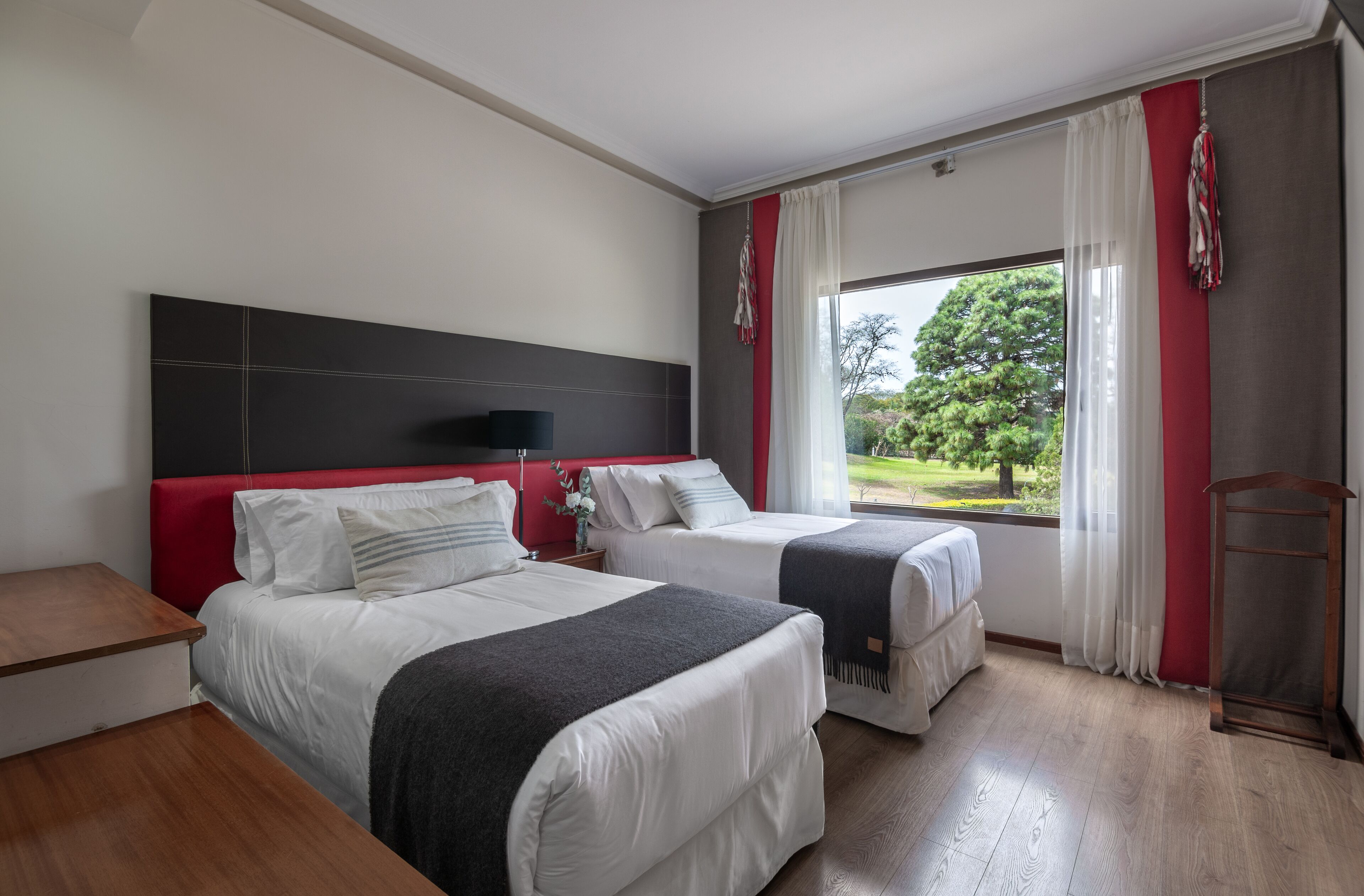A hotel room with two beds, a wooden floor, a red headboard, and a window with curtains.