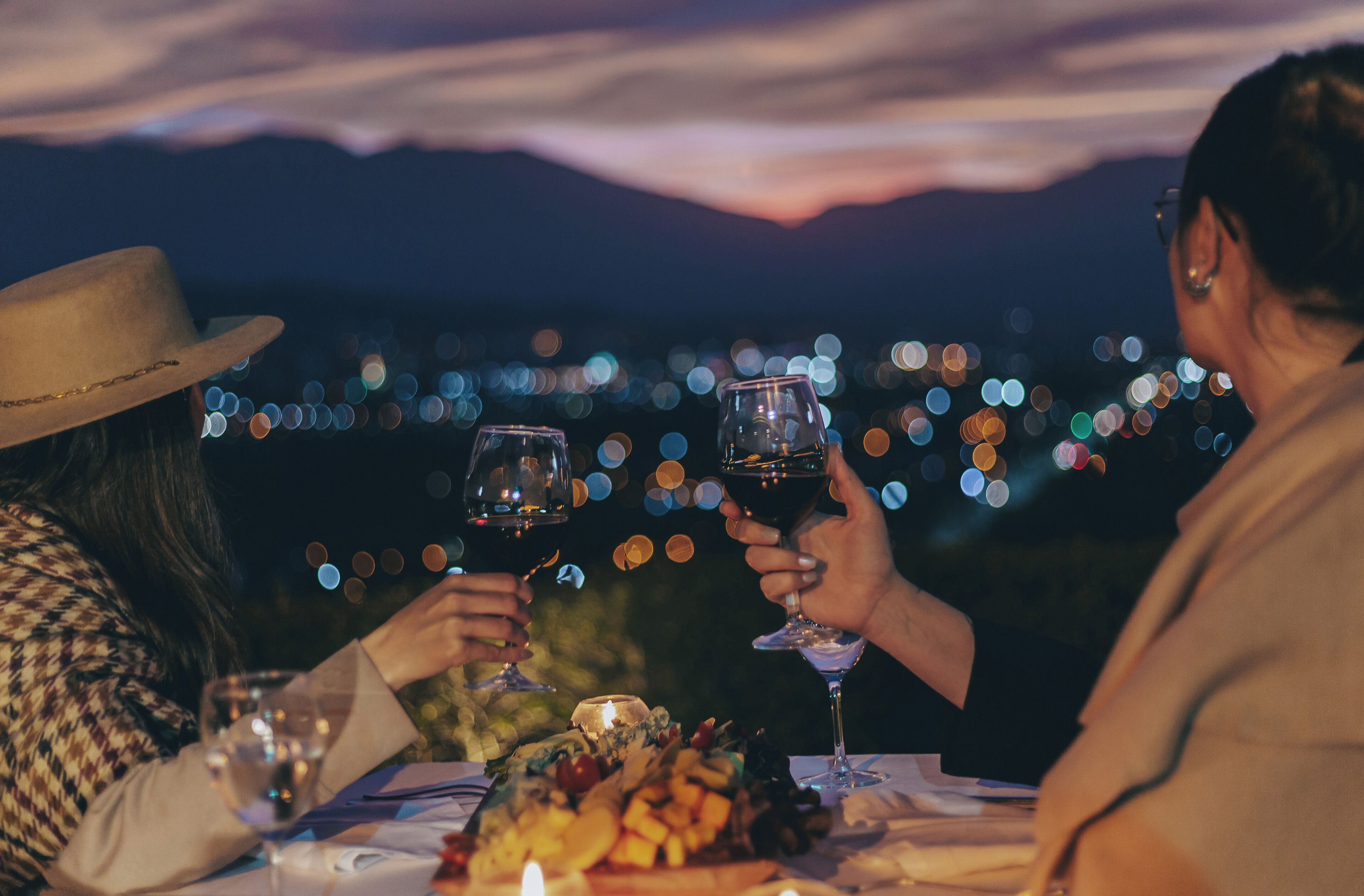 A couple toasting with wine glasses at a table with a fruit platter, set against a night sky with city lights.