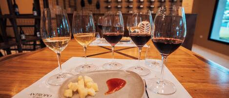 A wine tasting setup with glasses of white and red wine on a wooden table, a plate with cheese and jam, and a wine rack in the background.