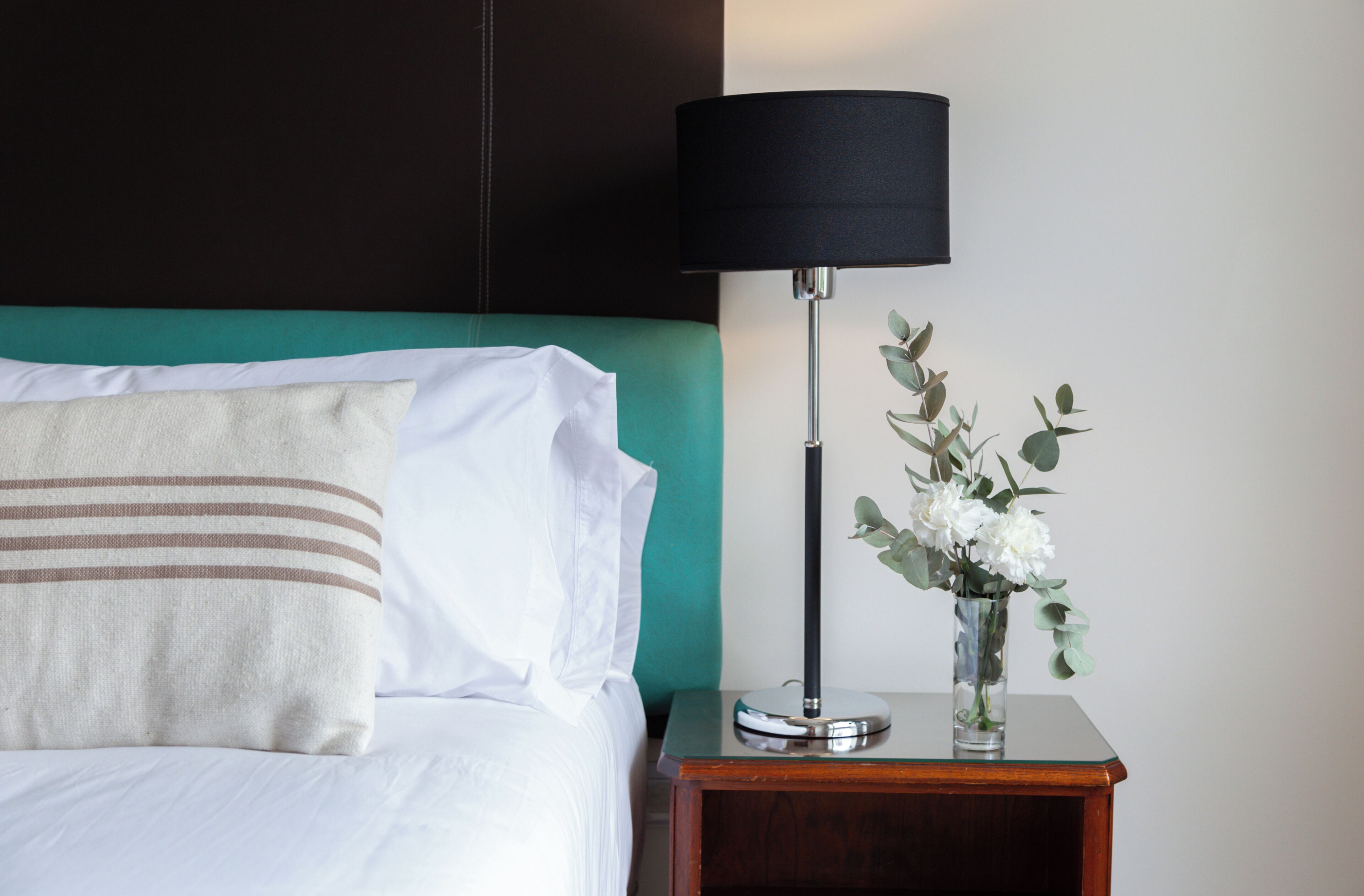 En pænt redt seng med hvide og stribede puder, en teal farvet sengegavl, en sort lampe og en vase med hvide blomster på et trænatbord.