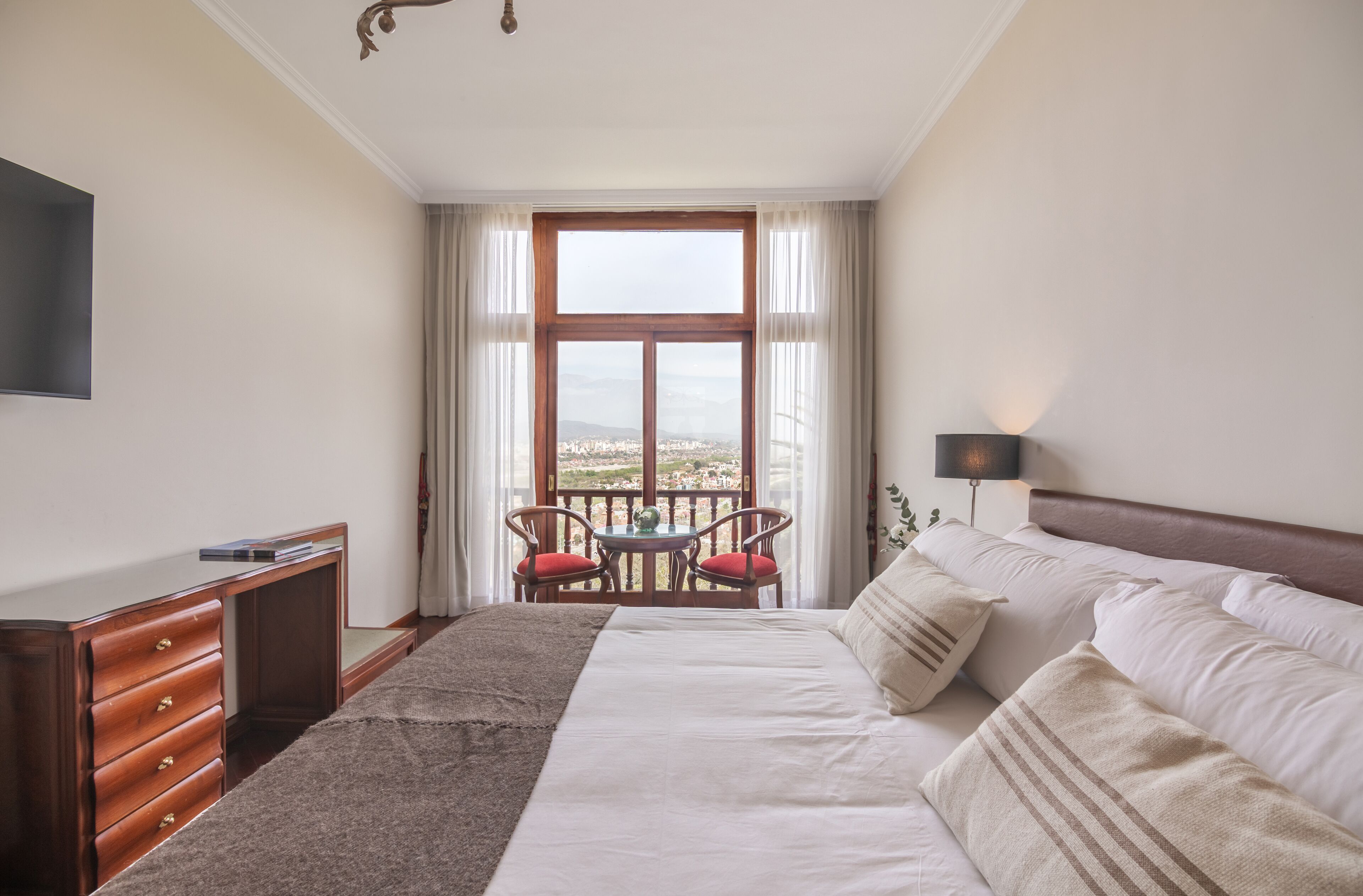 A hotel room with a large bed, a desk, a TV, a balcony with a table and chairs, and a view of the city.