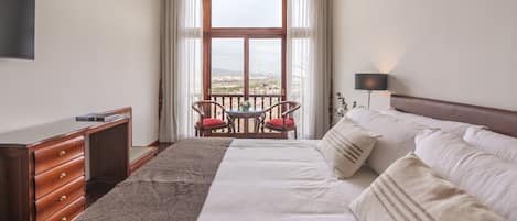 A hotel room with a large bed, a desk, a TV, a balcony with a table and chairs, and a view of the city.