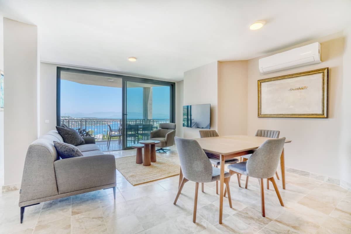 Uma sala de estar moderna com área de jantar, sofá, televisão e uma grande janela com vista para o mar.