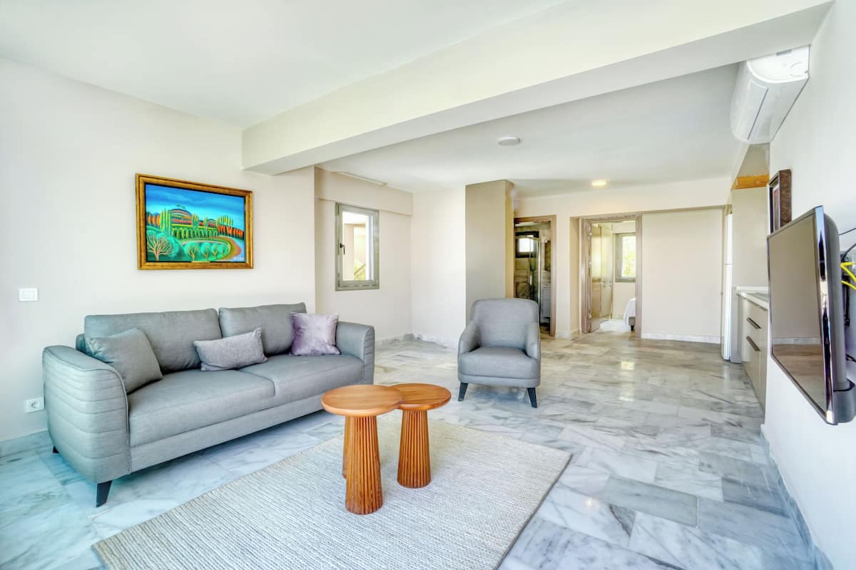 Uma sala de estar moderna com um sofá cinzento, duas mesinhas de cabeceira de madeira, uma poltrona cinzenta e um quadro na parede.