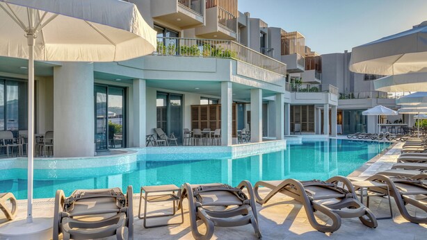 A poolside area with lounge chairs and umbrellas, a clear pool, and a modern building with balconies. - Olira Türkbükü Residences (Bodrum)