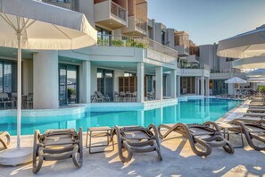 A poolside area with lounge chairs and umbrellas, a clear pool, and a modern building with balconies. - Olira Türkbükü Residences (Bodrum)