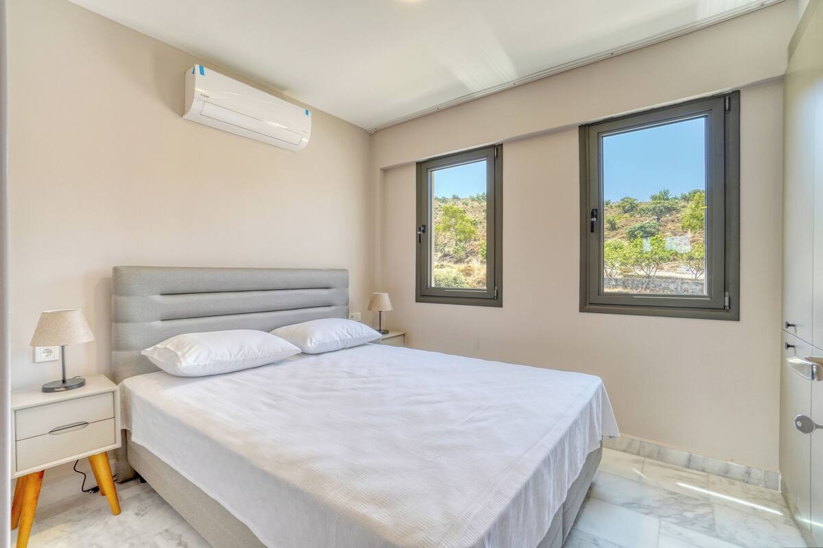 Quarto com cama, mesas de cabeceira, ar condicionado e duas janelas com vista para o exterior.