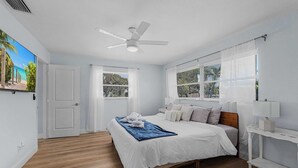 A bedroom with a bed, a ceiling fan, a window with blinds, a nightstand, and a wall-mounted TV.