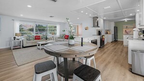 A modern kitchen with white cabinets, a dining area with a round table, and a living room with a sofa and a window.