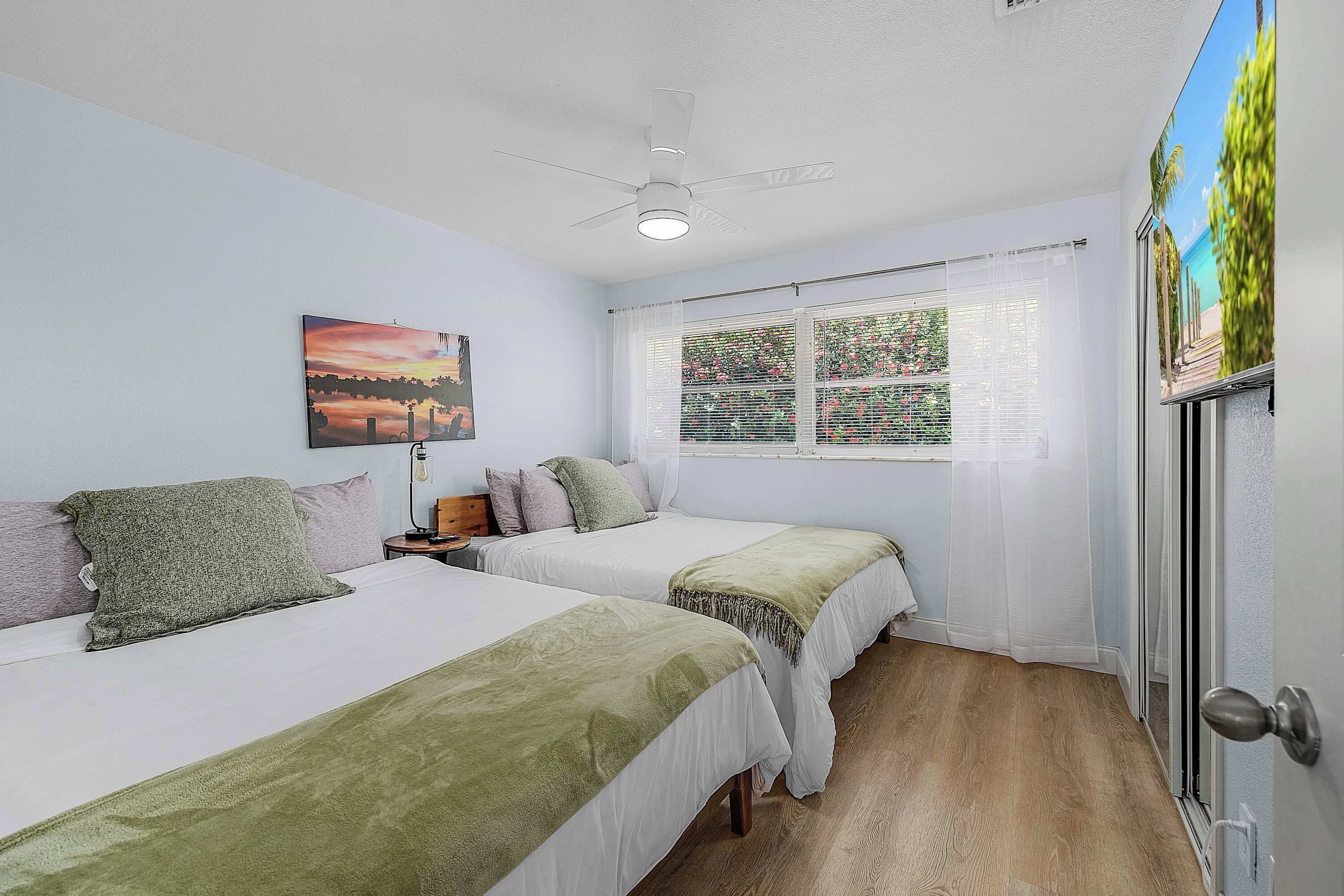 Ein Zimmer mit zwei Betten, einem Deckenventilator, einem Fenster mit Jalousien und einer Tür mit Griff.