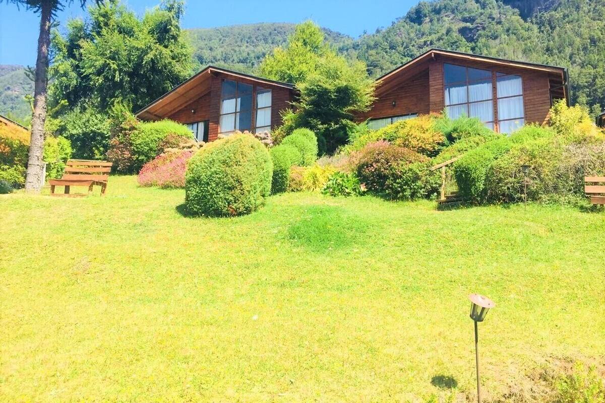 Two wooden cabins with large windows, surrounded by lush greenery and hills in the background.