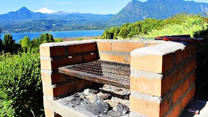 Una parrilla de ladrillo con rejilla metálica, rodeada de follaje y con vista a un lago con montañas al fondo.