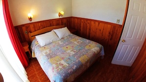 Un dormitorio con cabecera de madera, dos luces fijadas a la pared, una cama con colcha estampada y una puerta a la derecha.