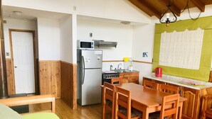 Cocina compacta con refrigerador, microondas y estufa. Hay una mesa de comedor de madera con sillas y un sofá verde en la sala.